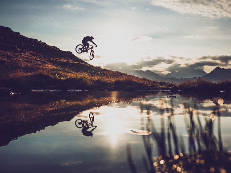 Ein Biker spiegelt sich beim Sprung im See, er ist auf einer Biketour auf Parsenn Gotschna. | © Davos Klosters Mountains